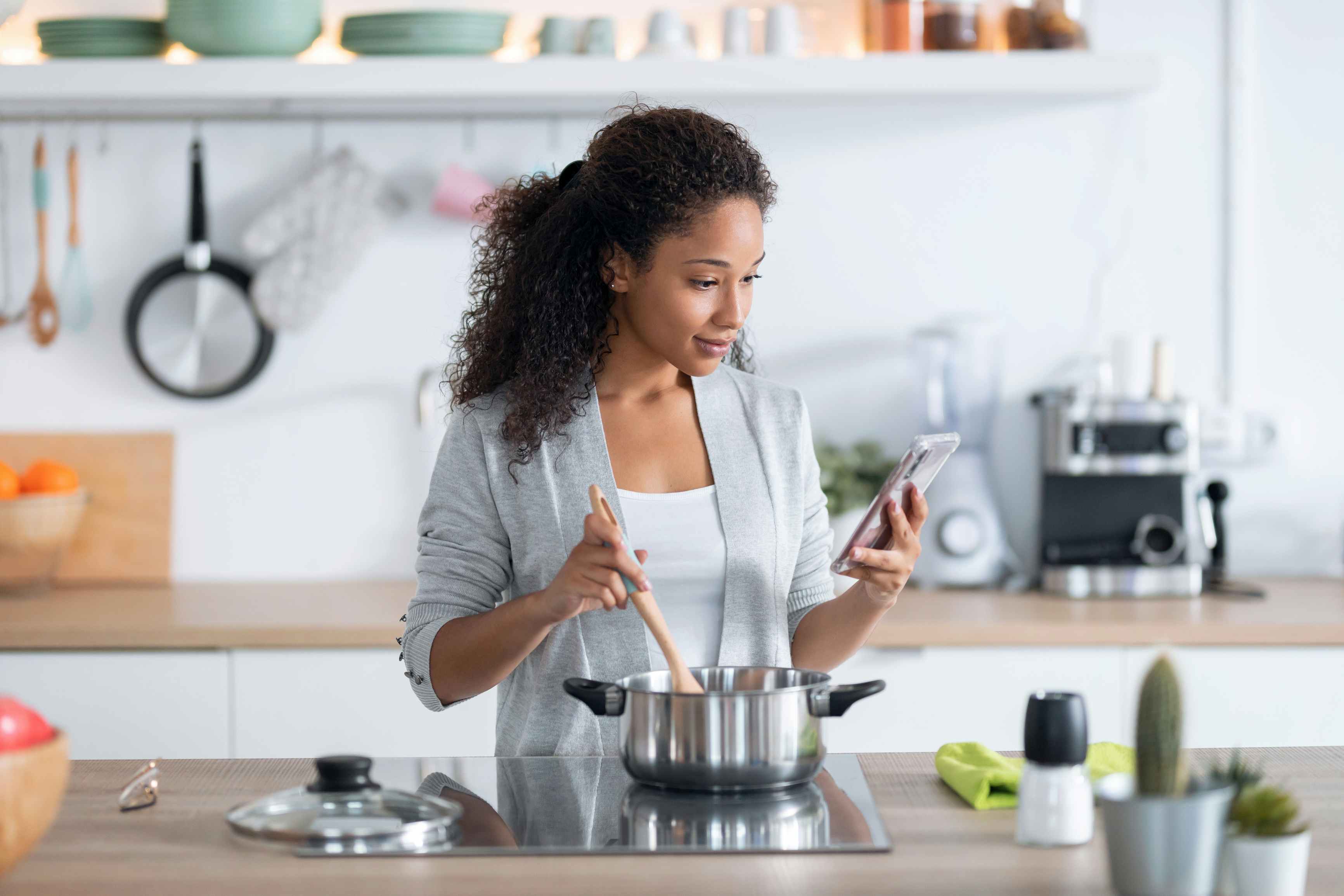 A woman cooking on a stove in a kitchen and looking at her phone