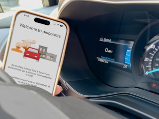 Someone holding up the discount page of the State Farm safe driving app next to an Odometer in a vehicle with very few miles on it