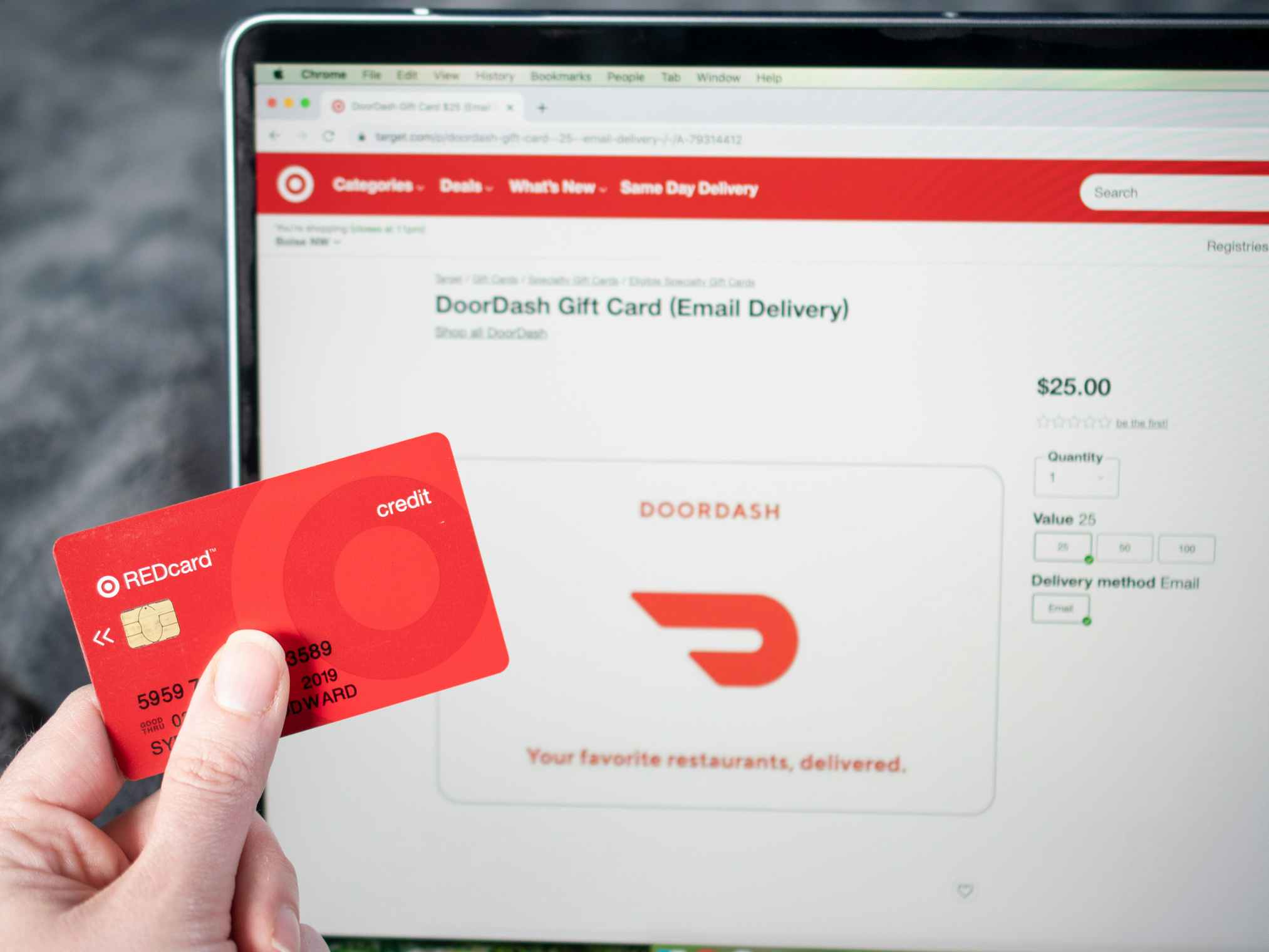 A hand holding a target red card in front of a computer screen showing DoorDash email gift cards on Target.com