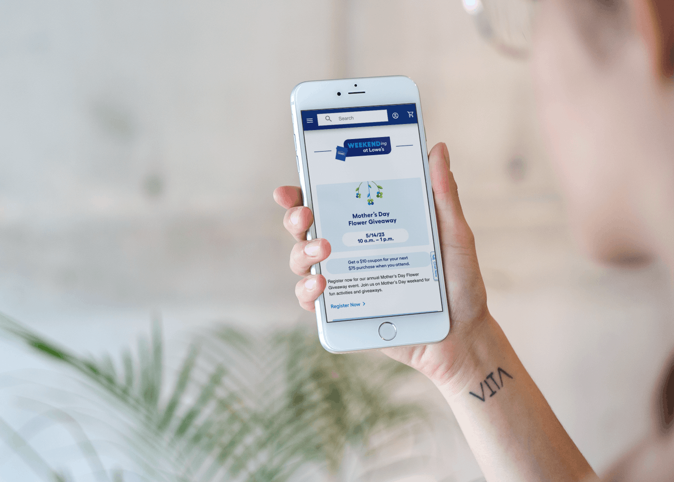 A woman holding up her phone and looking at the Lowe's event page online, showing a $10 coupon is available if you attend the Mother's Day Flower Giveaway on May 14, 2023
