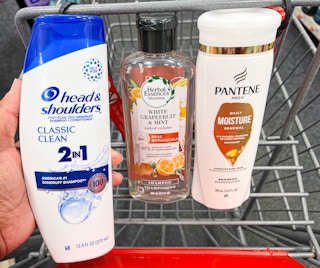 hand holding one bottle of head & shoulders shampoo, herbal essences shampoo, and Pantene shampoo in shopping cart