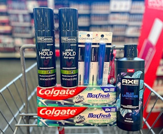 hand holding two Covergirl mascaras in front of one Axe body wash, two Tresemme hair sprays, and two Colgate Max Fresh toothpaste inside CVS shopping cart