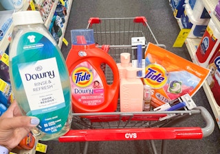 hand holding one bottle of Downy Rinse and Refresh in front of shopping cart with bottle of Tide liquid detergent, Monday shampoo and conditioner, one pack of Tide Pods, two Covergirl mascara packs, and one Revlon foundation inside