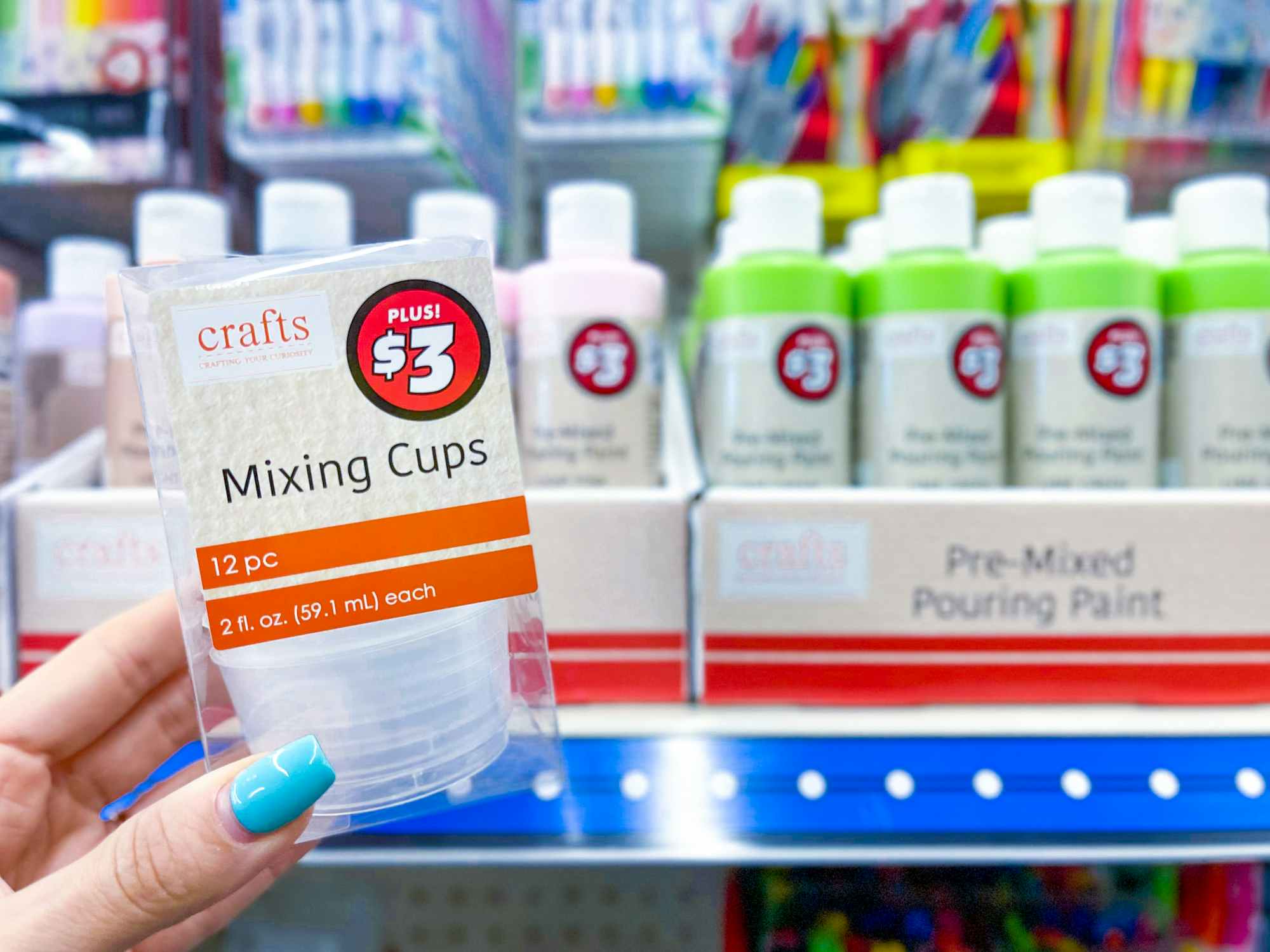 Paint mixing cups with a $3 price tag being held in front of some paint supplies on a shelf in the Dollar Tree Plus section