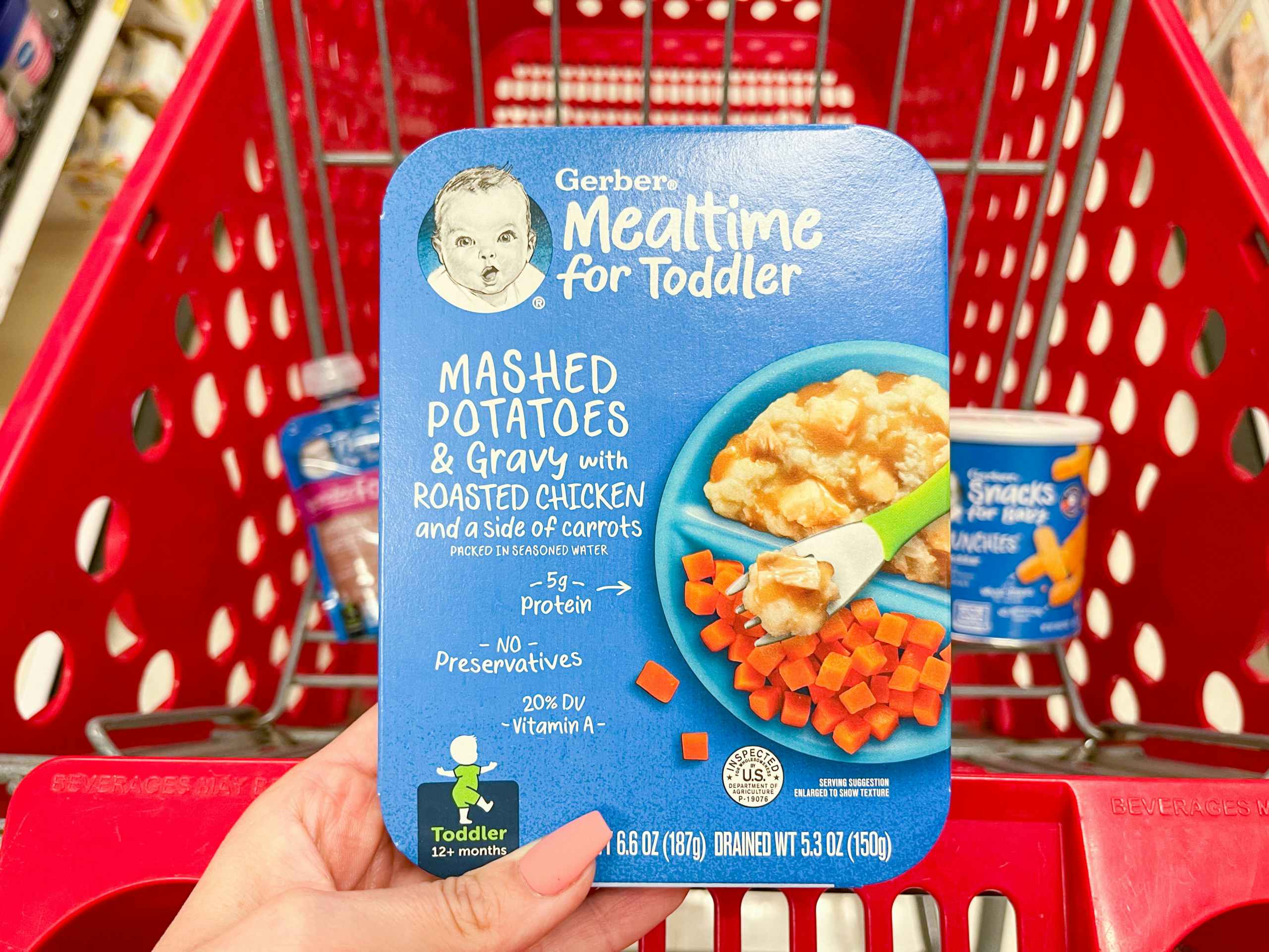A Gerber Mealtime for toddler meal held out by hand in front of a store cart with other Gerber items in it.