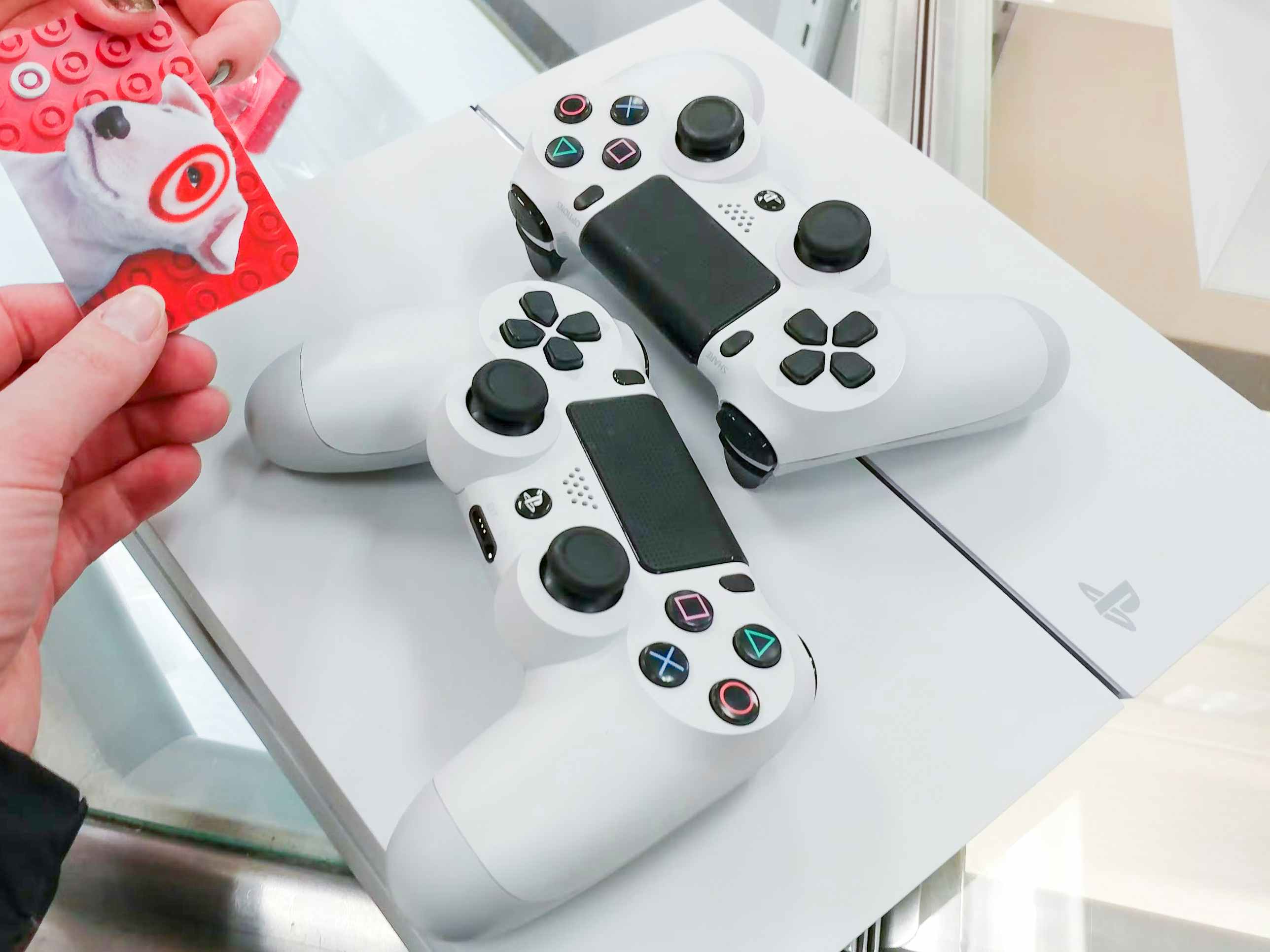 An old PlayStation gaming console and two controllers on the counter with a hand holding a target gift card