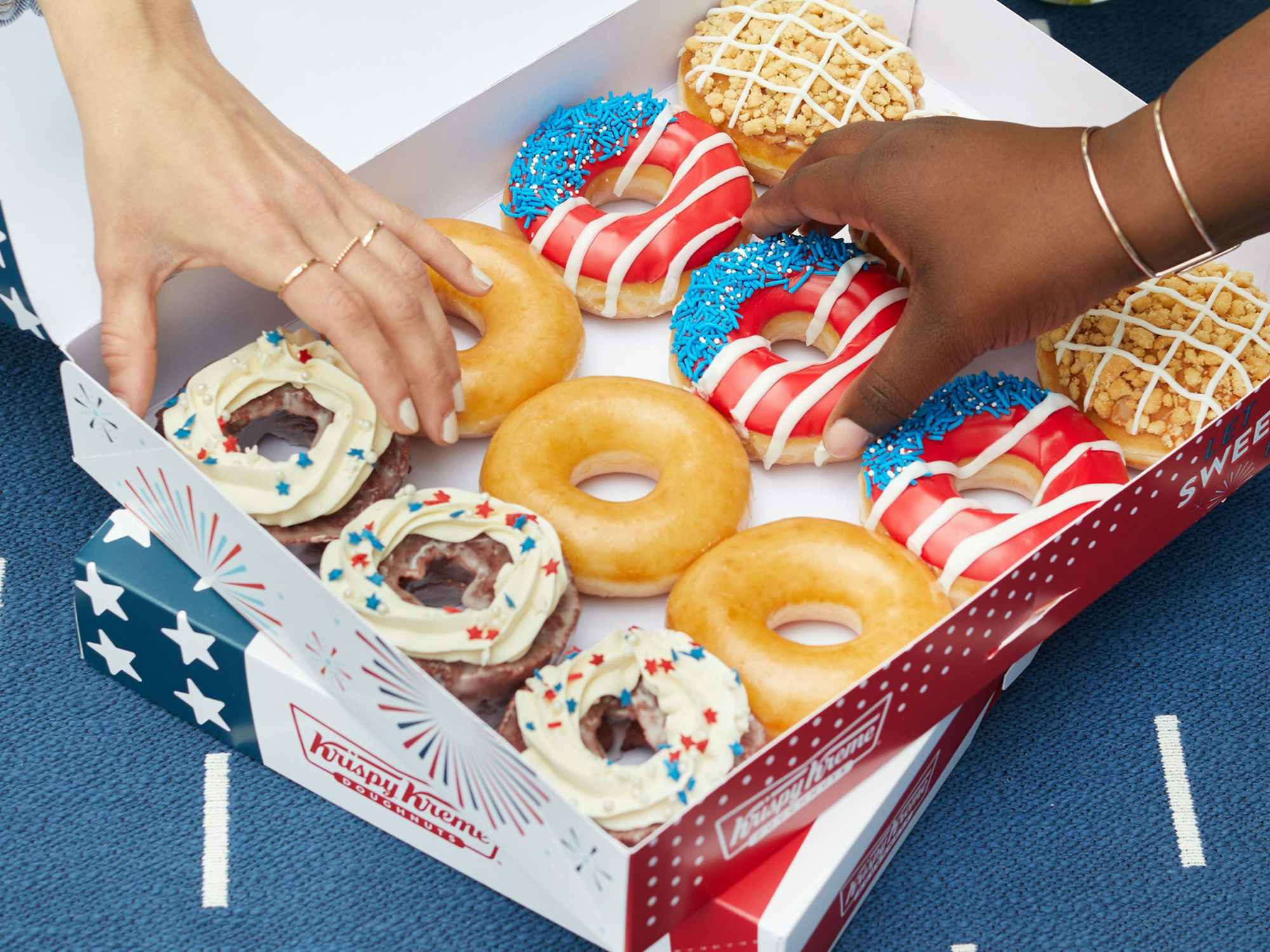 People reaching to take some Red, White, and Blue themed doughnuts from a Krispy Kreme box