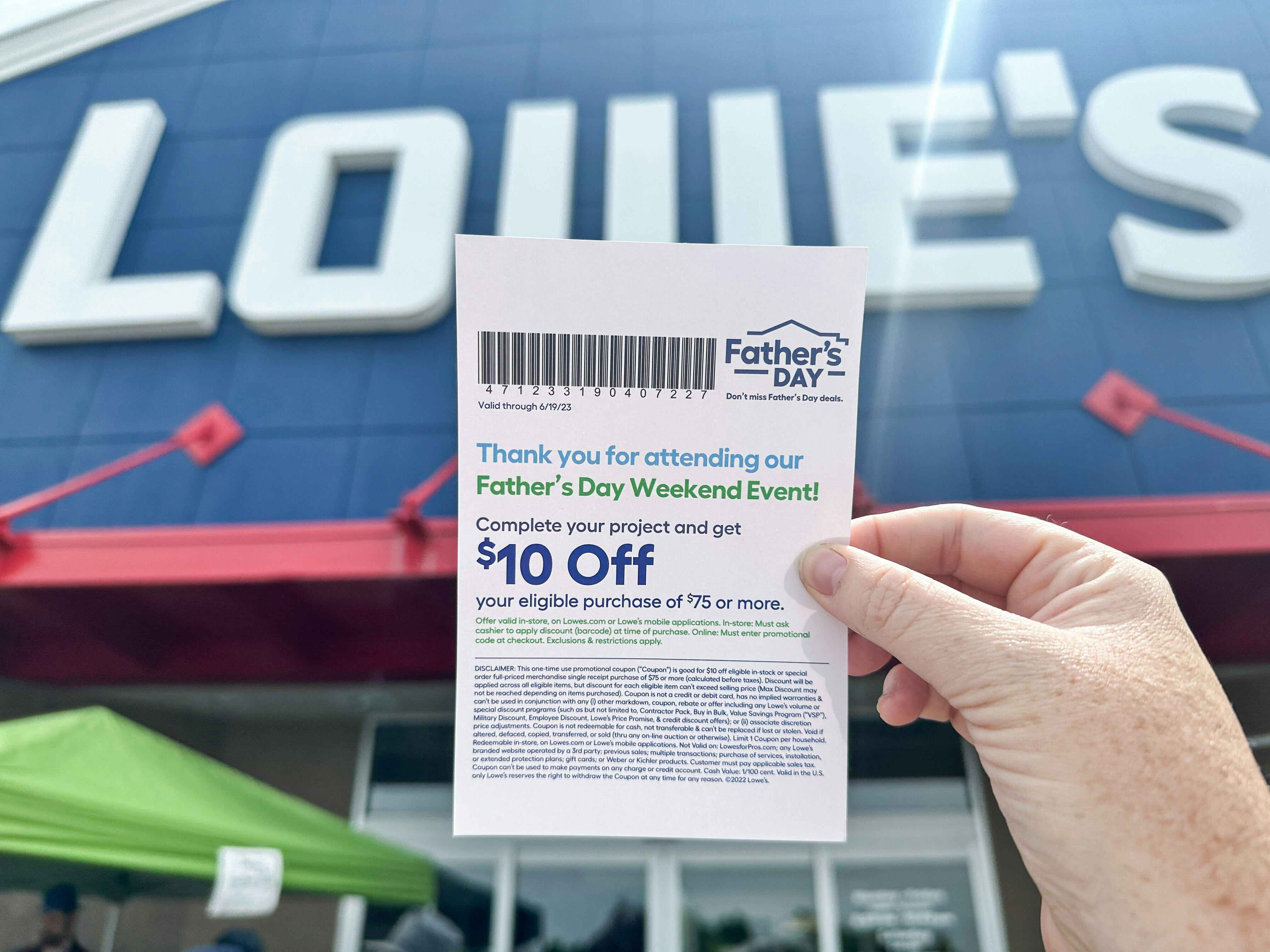 A woman's hand holding up a $10 off coupon in feront of the Lowe's storefront sign