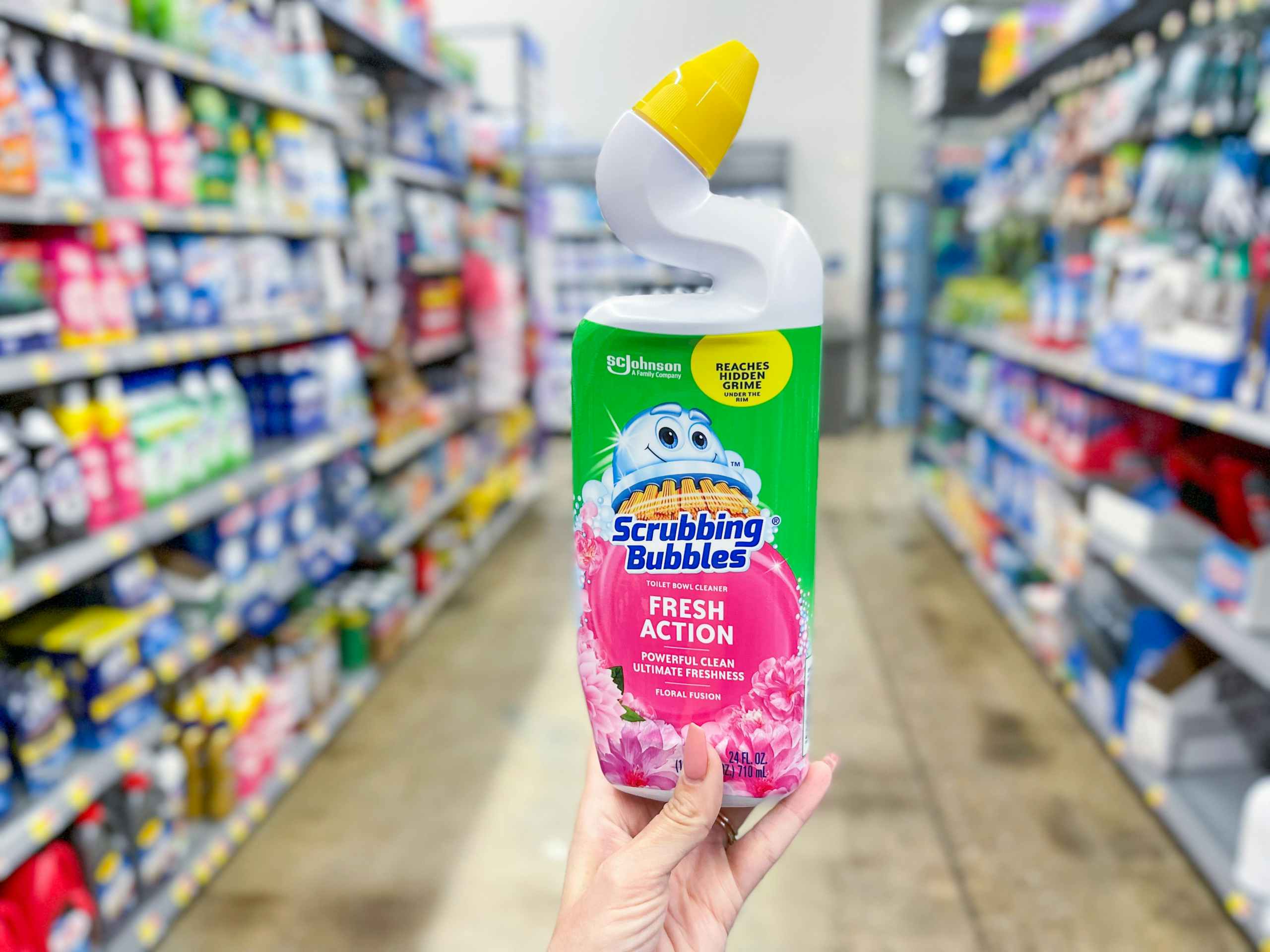 A bottle of scrubbing bubbles fresh action held out by hand in front of a cleaning aisle in a store.