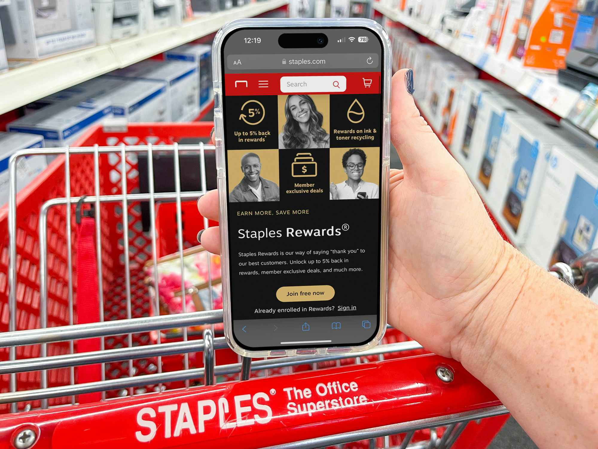 A person's hand resting on the handle of a Staples shopping cart, holding a cell phone displaying the Staples Rewards page on their website.