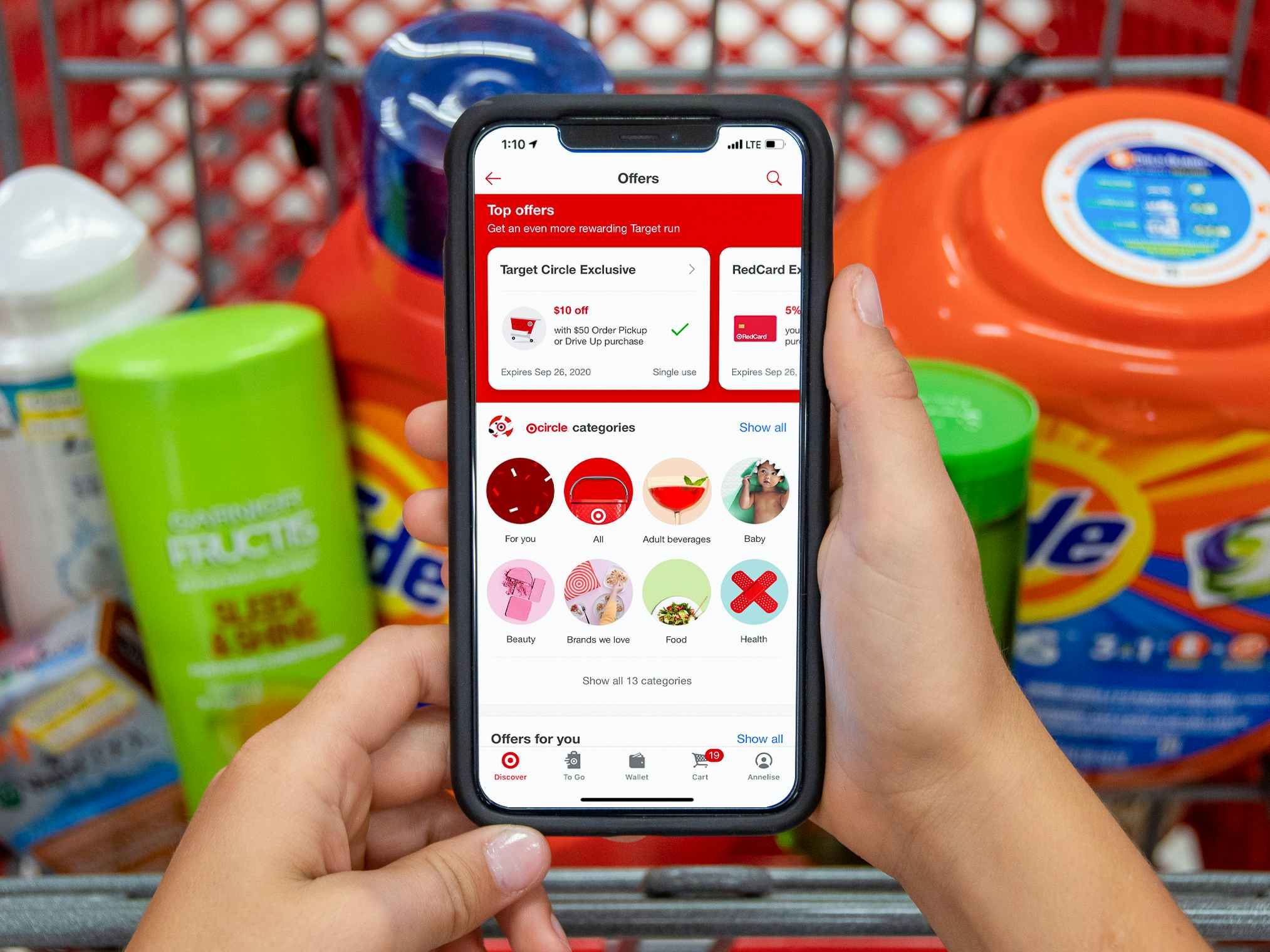 A person's hands holding a phone displaying the Target mobile app's Offers page in front of a Target shopping cart's basket filled with products.