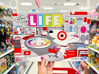 a woman's hand holding up the target game of life board game in front of a red shopping cart in the store