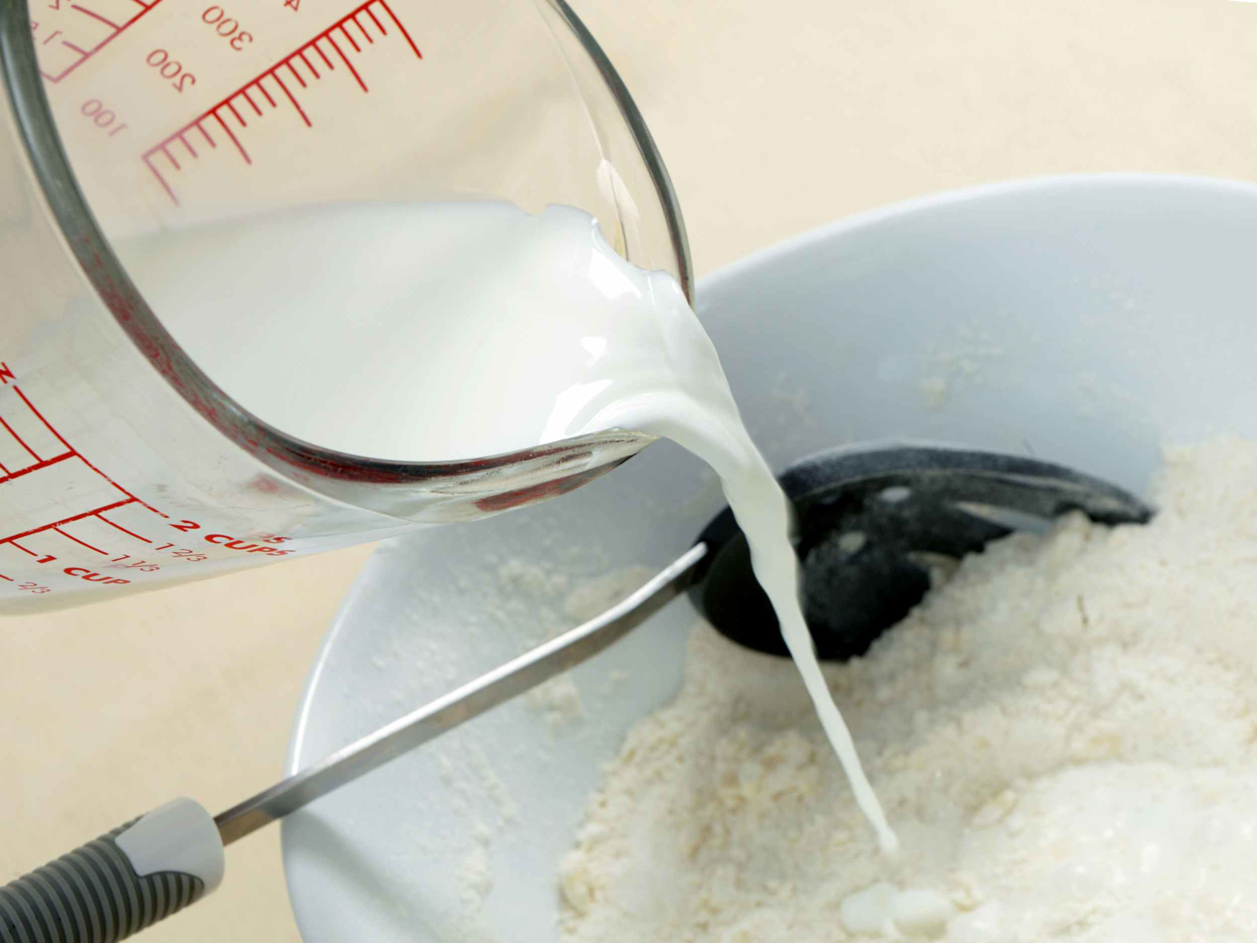 pouring milk from a measuring cup into a bowl with a spoon and flour