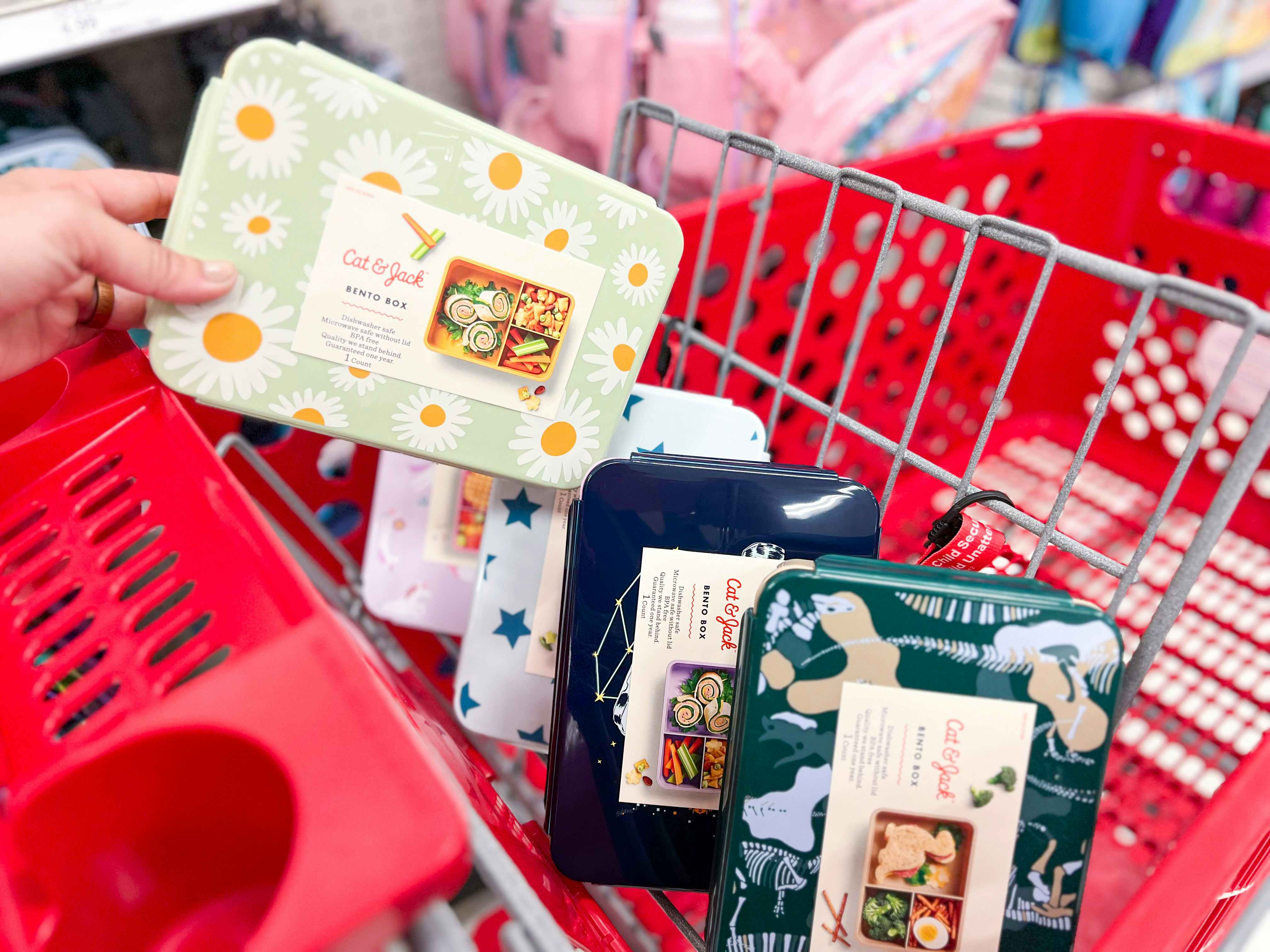 a woman's hand putting a target cat and jack bento box with daisies on it into her shopping cart with three other bento boxes in the cart already
