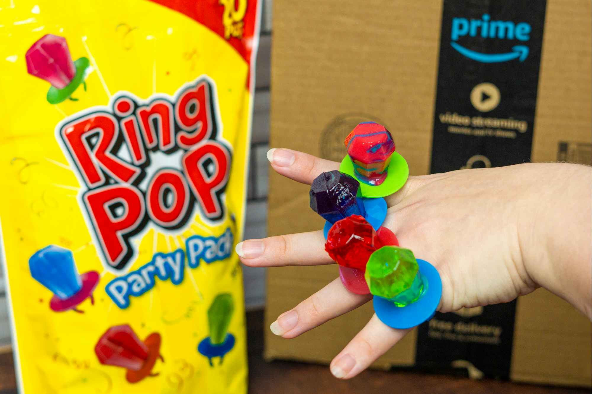 someone holding up their hand, a ring pop on each finger, in front of the Ring Pop party pack bag and an amazon prime box