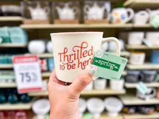 hand holding up a cream mug that says "thrilled to be here" in red cursive writing