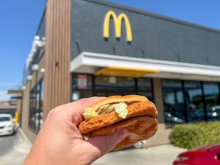 someone holding up a Grand McChicken sandwich in front of a McDonald's